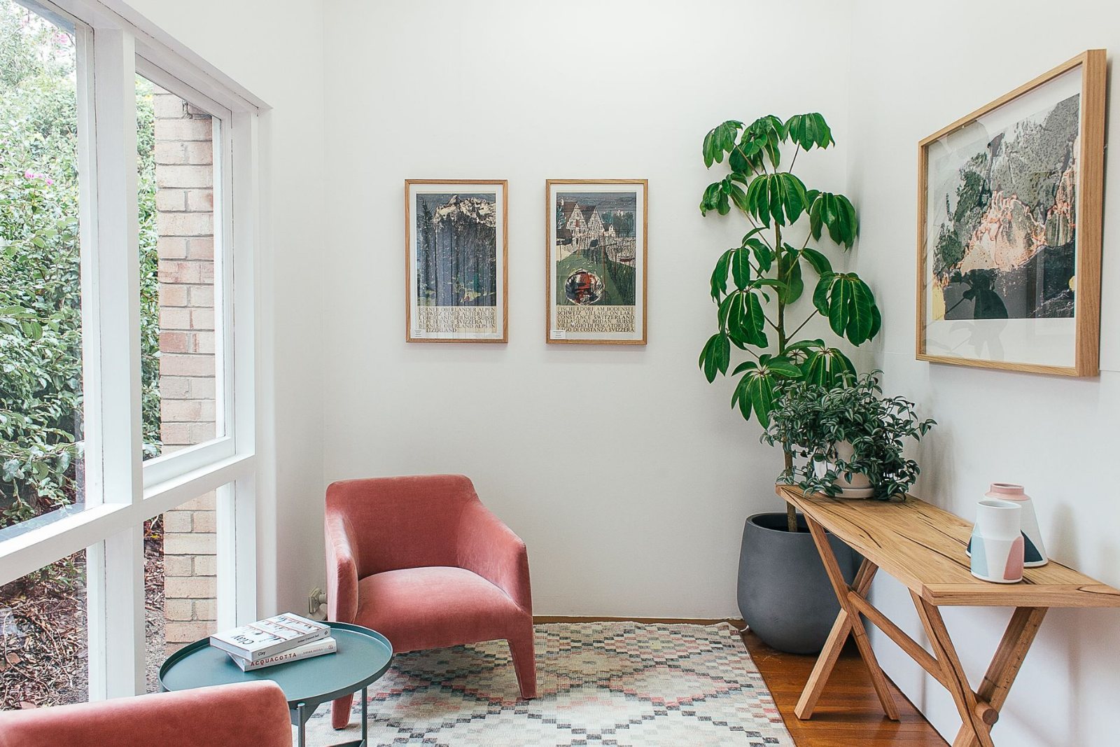 A lived-in living room after the work is finished: folded throw blankets squared on the sofa arm, cushions straightened, a trailing plant on the windowsill, a book and one mug on a low wooden table, late-morning light raking across a clean floor